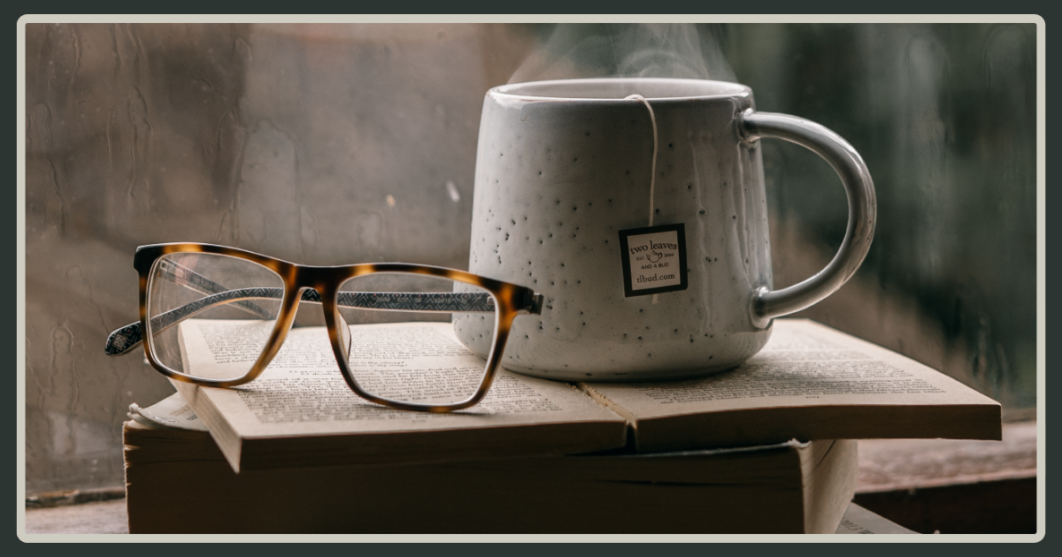 A pair of tortoiseshell glasses resting on an open book next to a steaming ceramic mug of tea, set against a window with raindrops.