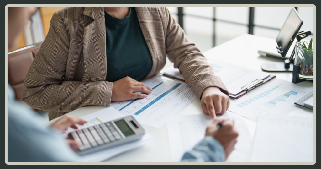 Two professionals sitting at a desk reviewing printed data charts and financial spreadsheets, with one person using a calculator.