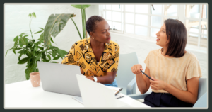 Two women sitting at a white desk with laptops, engaged in an earnest and casual face-to-face conversation.