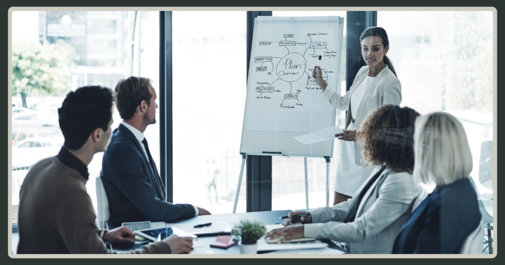 A female leader standing at a whiteboard, pointing to a "Plan Overview" diagram while presenting to a team in a modern boardroom.