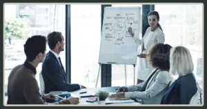 A female leader standing at a whiteboard, pointing to a "Plan Overview" diagram while presenting to a team in a modern boardroom.