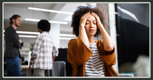 A woman at her desk holding her head in her hands in frustration while colleagues talk in the background of a busy office.