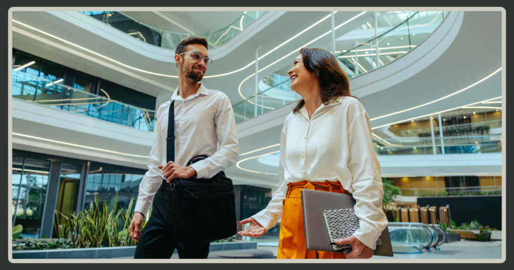 A man and a woman walking and talking through a bright, multi-level modern office atrium while carrying work bags and a laptop.