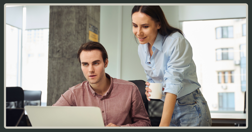A woman in a light blue shirt leans over a male colleague's shoulder to review work on a laptop screen in a modern office.