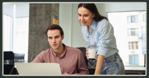 A woman in a light blue shirt leans over a male colleague's shoulder to review work on a laptop screen in a modern office.