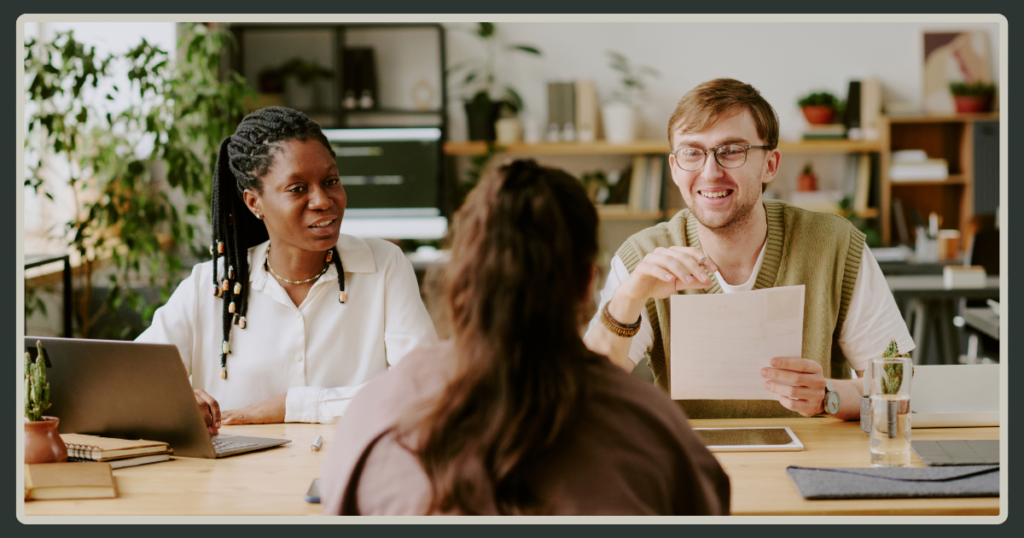 Two smiling interviewers, a Black woman and a man with glasses, review a candidate’s resume during a professional job interview.