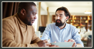 A Black man in a tan sweater and a bearded man in a blue shirt sit close together, focused on a digital tablet during a discussion.