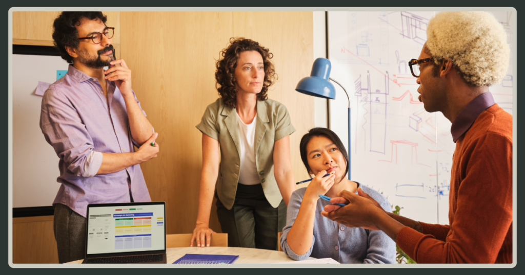 A diverse team of four colleagues engages in a brainstorming session around a table with a laptop and whiteboard in a bright office.