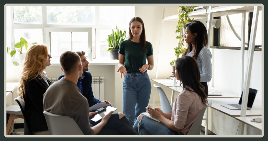 A young woman stands in the center of a bright, modern office, gesturing while leading a discussion with a small seated group of colleagues.