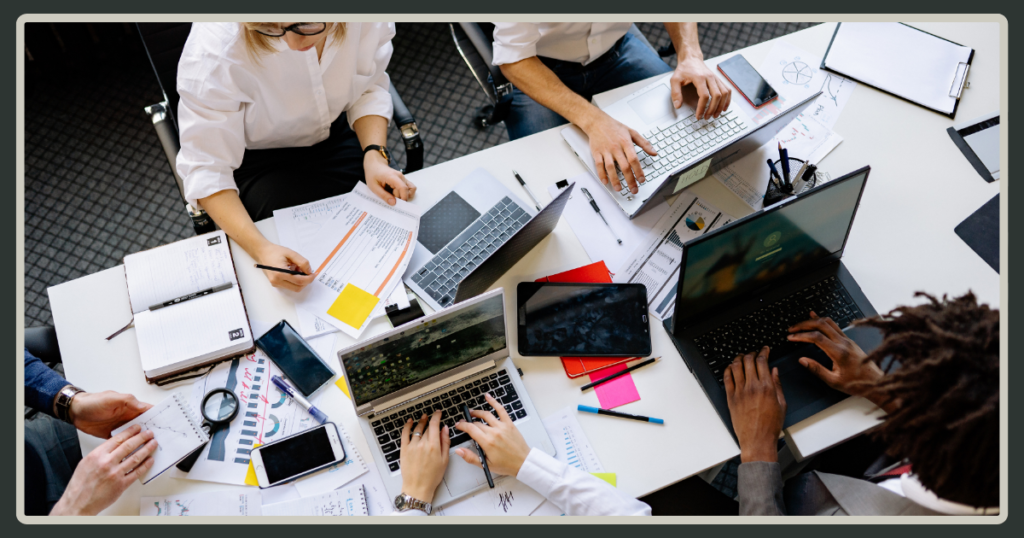 An overhead, "bird's-eye" view of a shared office table where multiple people are simultaneously working on laptops and notebooks.