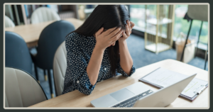 A woman in a polka-dot shirt sits at a desk with an open laptop, covering her face with her hands in a moment of distress or overwhelm.