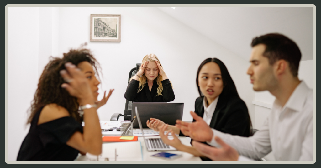 A frustrated manager holds her head in her hands in the background while three employees engage in a heated, gestural argument in the foreground.