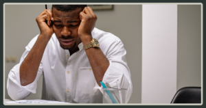 A Black man in a white dress shirt sitting at a desk, clutching his head in his hands with a look of deep stress or exhaustion.