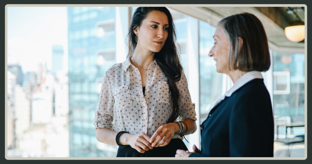 Two professional women engaged in a thoughtful, serious conversation in a bright office space with a city skyline in the background.