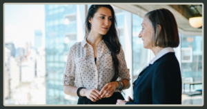 Two professional women engaged in a thoughtful, serious conversation in a bright office space with a city skyline in the background.