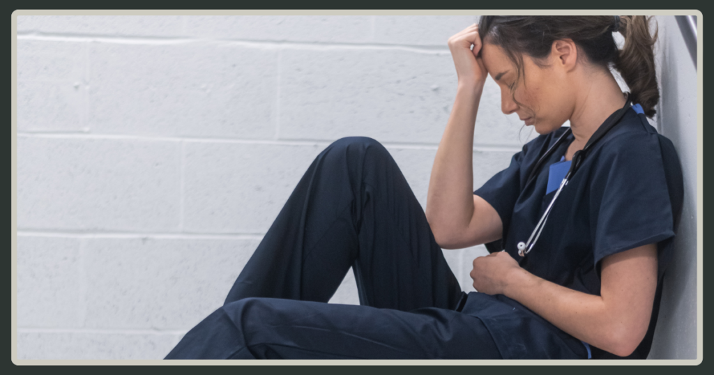 A woman in blue medical scrubs sitting on the floor against a white brick wall, resting her head in her hand in a moment of grief or fatigue.