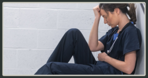 A woman in blue medical scrubs sitting on the floor against a white brick wall, resting her head in her hand in a moment of grief or fatigue.
