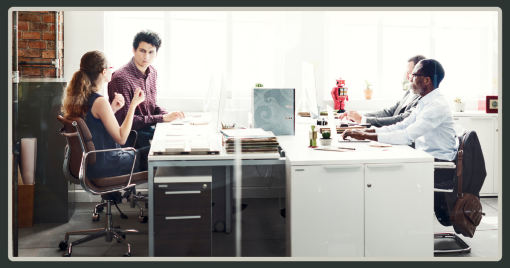 A view of a modern open-plan office where colleagues work at long shared desks; one woman is gesturing while talking to a coworker.