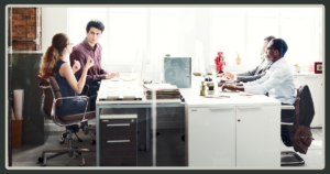 A view of a modern open-plan office where colleagues work at long shared desks; one woman is gesturing while talking to a coworker.
