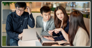 Four Asian professionals huddled around a small table, focused intently on a laptop and tablet during a collaborative discussion.