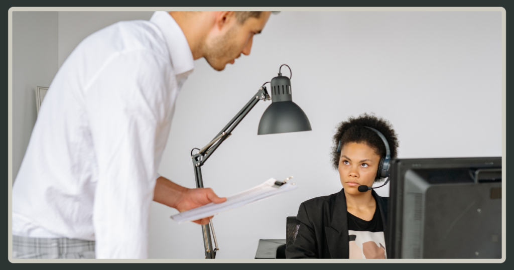 A man in a white shirt holding a clipboard stands over a female employee at her desk, who is wearing a headset and looking up at him during a serious talk.