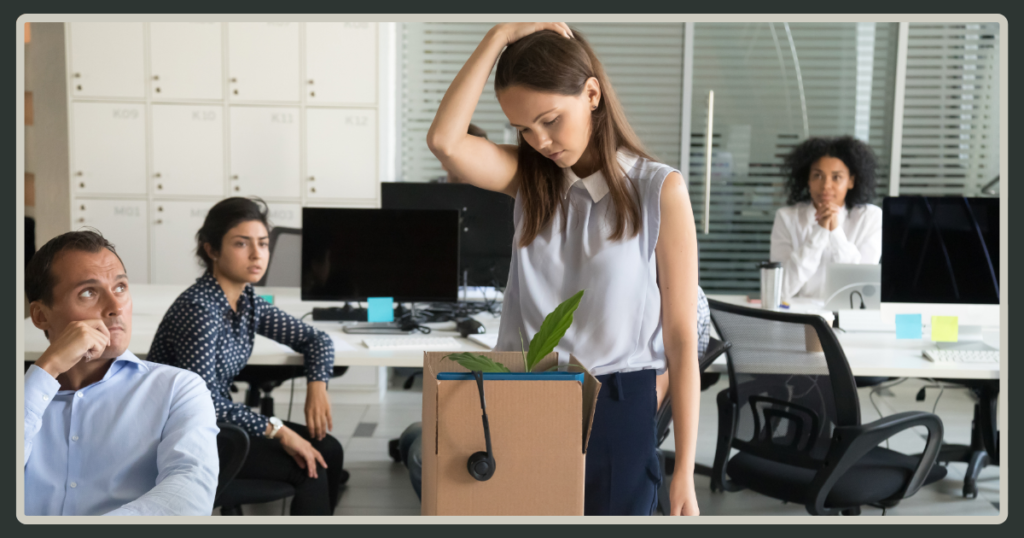 A young woman stands in the center of an office looking down sadly, holding a cardboard box filled with her desk items while colleagues watch from the background.