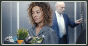 A somber woman with curly hair carries a box of her personal office belongings, including a plant and a clock, while a man in a suit stands by an elevator in the background.