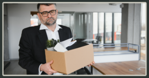 A dejected older man with glasses and a beard holds a cardboard box of office supplies while standing in a modern, open office space.