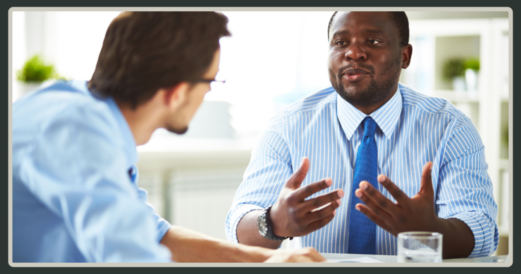 A man in a blue striped shirt and tie gestures with both hands while engaged in an earnest face-to-face discussion with a colleague.