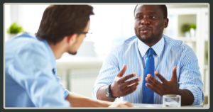 A man in a blue striped shirt and tie gestures with both hands while engaged in an earnest face-to-face discussion with a colleague.