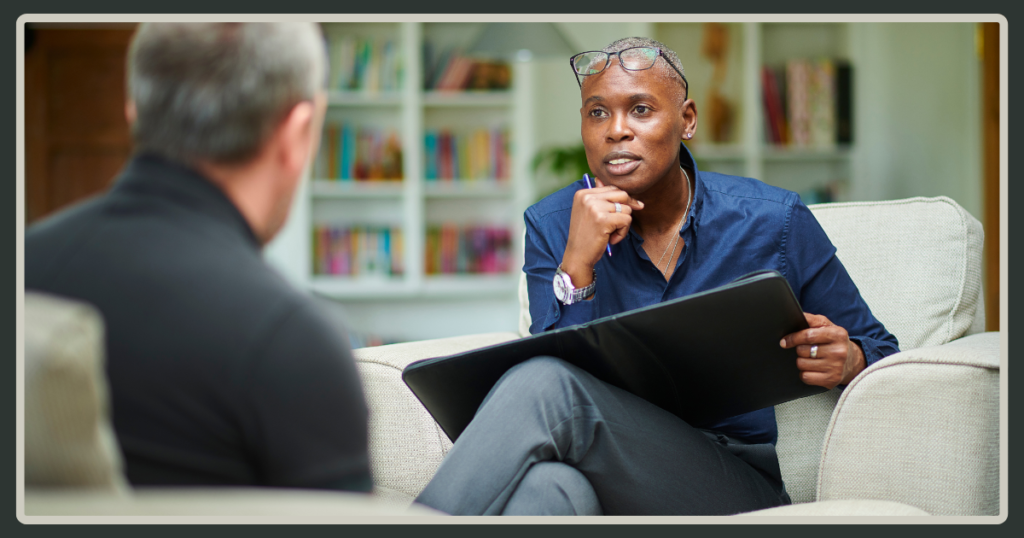 A professional Black woman with glasses sits in a neutral-toned armchair, listening intently and holding a portfolio during a one-on-one session.