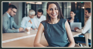 A woman in a gray top smiles warmly at the camera in the foreground, while a diverse team of professionals works at a conference table in the background.