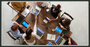 An overhead view of a diverse group of people sitting around a wooden table with laptops, tablets, and coffee, with two individuals shaking hands.