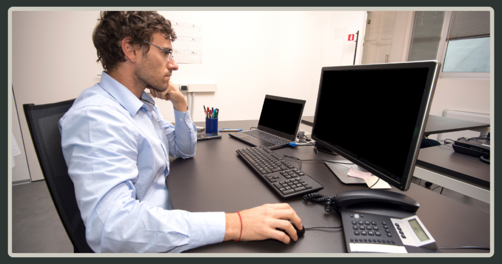 A man with glasses sits alone at a desk with multiple monitors and an office phone, appearing thoughtful and slightly overwhelmed while staring at a screen.