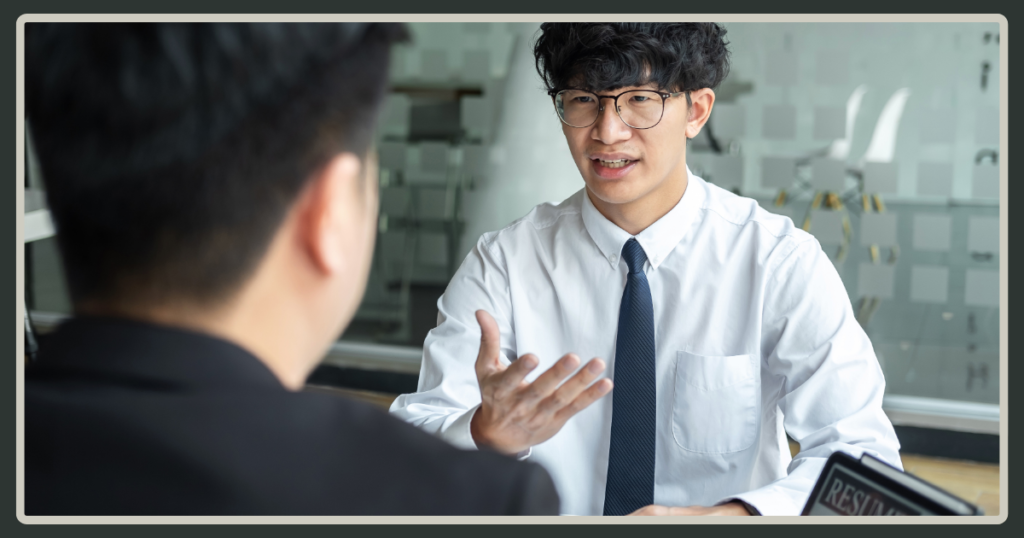 A young man with glasses and a tie gestures expressively while answering questions during a formal job interview.