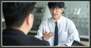 A young man with glasses and a tie gestures expressively while answering questions during a formal job interview.