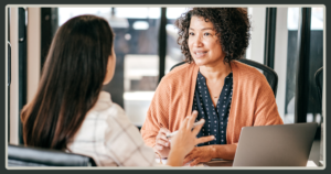 A woman with curly dark hair wearing an orange cardigan engages in a friendly professional conversation with a candidate or colleague across a table.