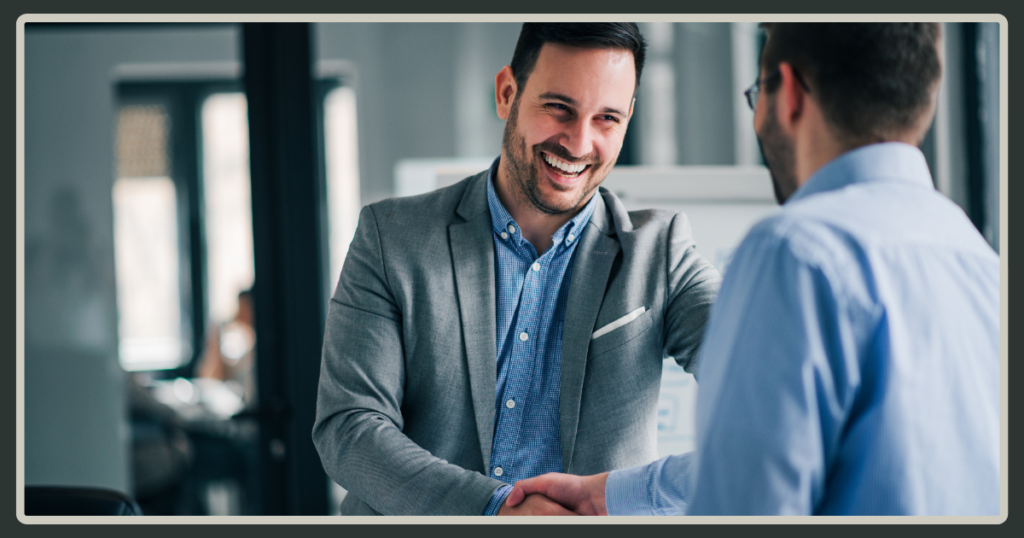 A smiling man in a gray blazer shakes hands with a colleague, signifying a warm welcome or a successful agreement in a professional office setting.
