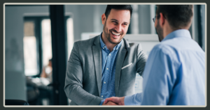 A smiling man in a gray blazer shakes hands with a colleague, signifying a warm welcome or a successful agreement in a professional office setting.