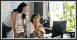 Two women in a professional office environment work together at a desk, looking intently at a computer monitor while discussing a project.