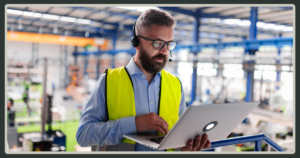 A man with a beard and glasses wearing a high-visibility safety vest and a headset works on a laptop in a large industrial or warehouse facility.