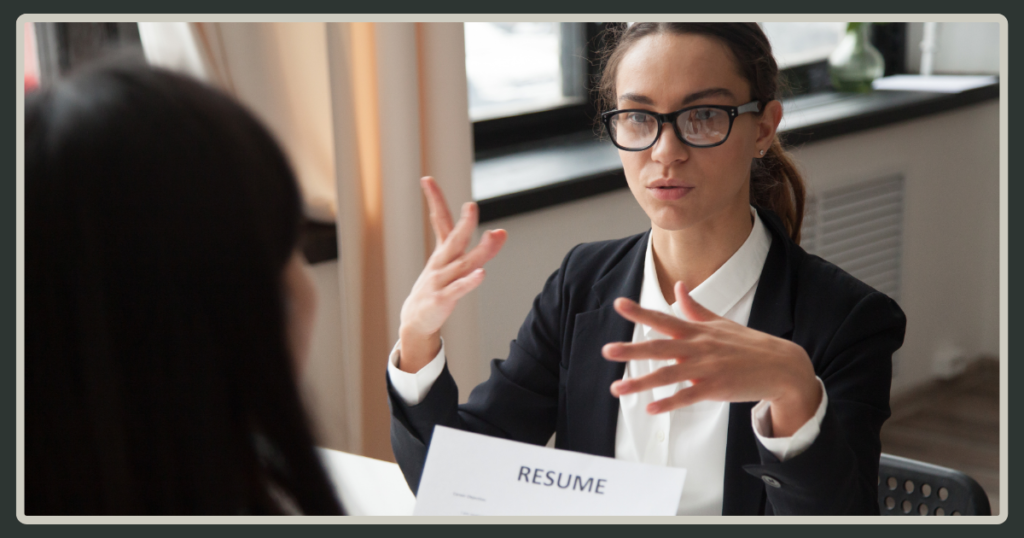A woman with glasses and a black blazer gestures with her hands while conducting an interview, with a "RESUME" document visible on the table.