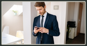 A man in a dark suit and tie leans against an office pillar while focused on his smartphone, reflecting a moment of quiet communication or isolation.