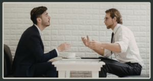 Two men in professional attire sitting at a small table, engaged in an earnest, hand-gestured conversation against a white brick wall.