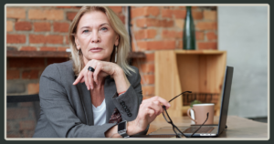 A serious-looking senior female executive in a gray blazer resting her chin on her hand at a desk with a laptop and a brick wall background.