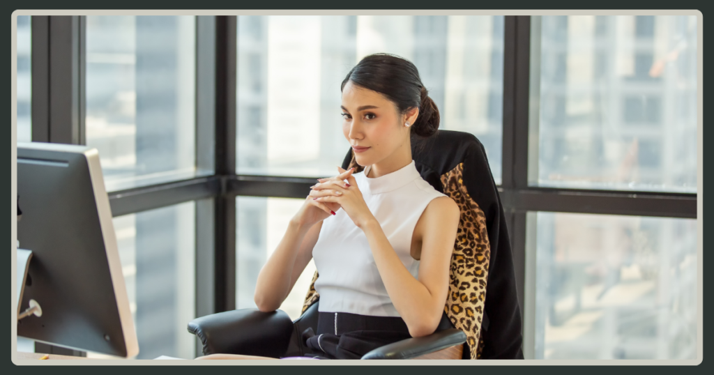A professional woman in a white top sitting in a high-back office chair with a leopard-print jacket, looking thoughtfully toward an office window.