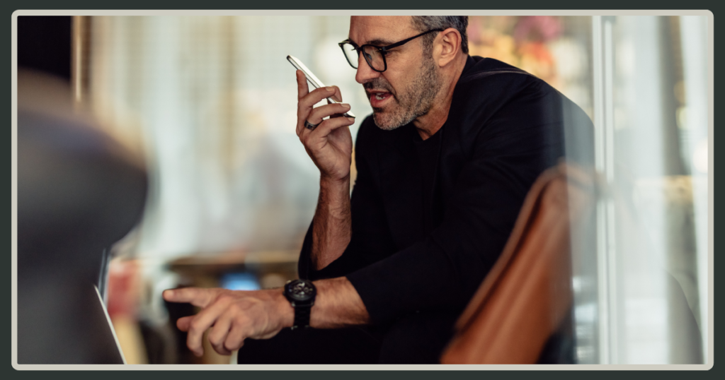 A male leader with glasses speaking into a smartphone while pointing at a laptop screen in a blurred, modern office setting.
