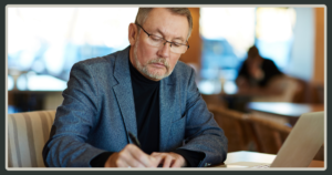 A senior man with glasses and a gray suit jacket looking down and writing on a document while sitting at a desk with a laptop.