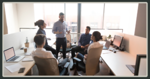 A male manager standing and gesturing while leading an informal team meeting with four colleagues in a bright, open office workspace.
