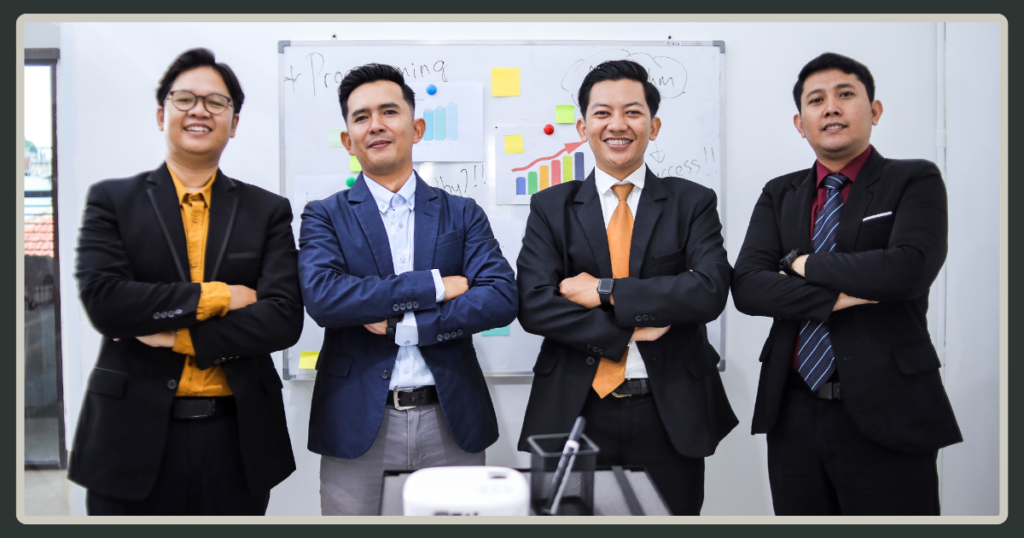 Four male professionals in blazers standing together with crossed arms and confident smiles in front of a whiteboard with charts.